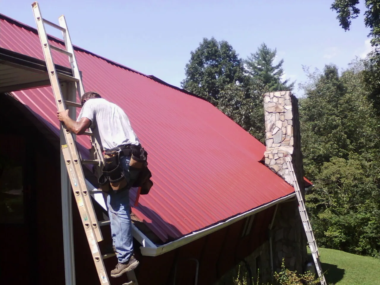 Roofer on a ladder beside a red metal roof and stone chimney — Soffit & Fascia Repair by Roofing Brothers in Elk